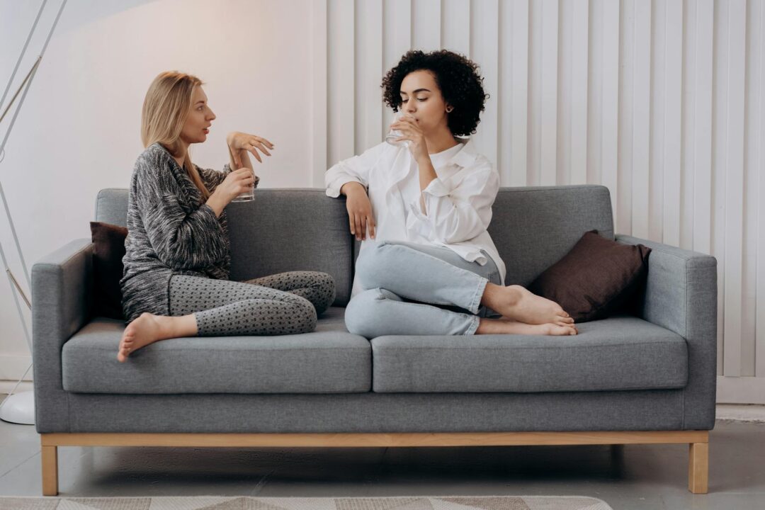 two women sitting on a gray couch drinking water