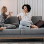 two women sitting on a gray couch drinking water