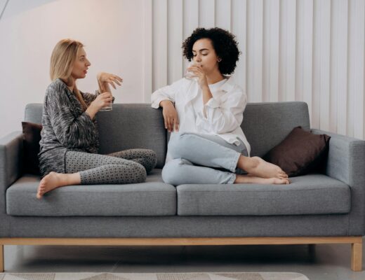 two women sitting on a gray couch drinking water